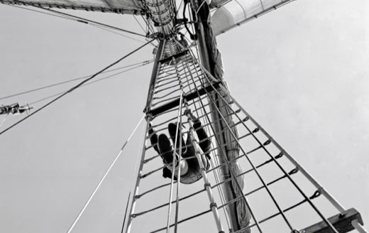 View from below of a person climbing a ship's mast with rigging and sails visible.