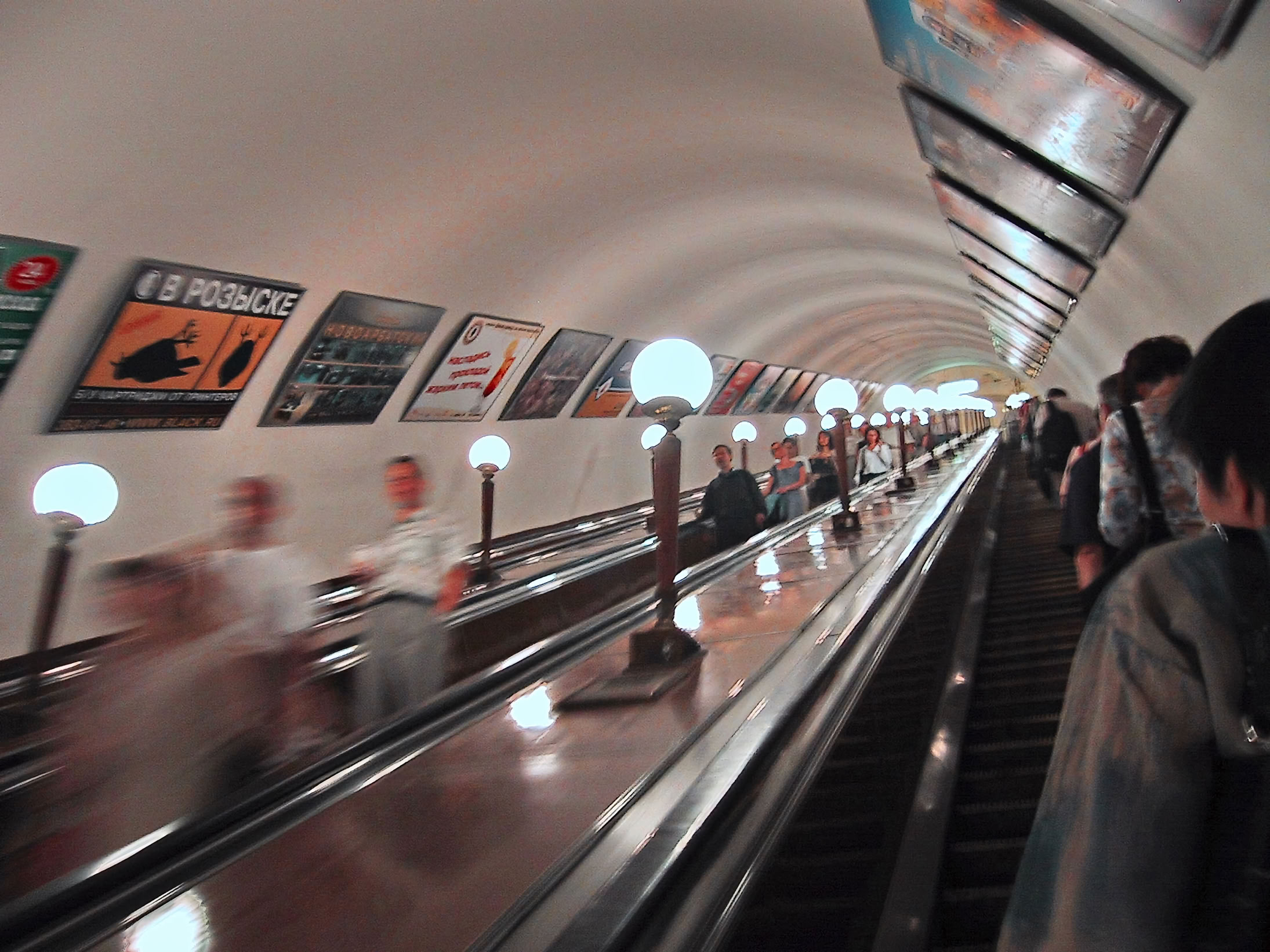 A long underground escalator with people ascending and descending, surrounded by illuminated lamps and advertisements on the walls.