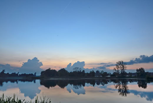 A serene lake reflecting the sky and clouds at sunset, with silhouettes of trees along the horizon.