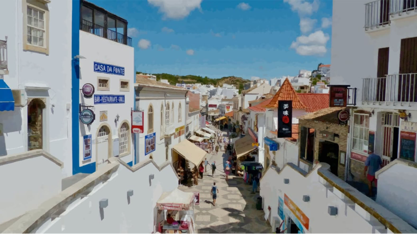 A narrow street lined with white buildings, some with blue accents, and a few people walking. The sky is blue with scattered clouds.
