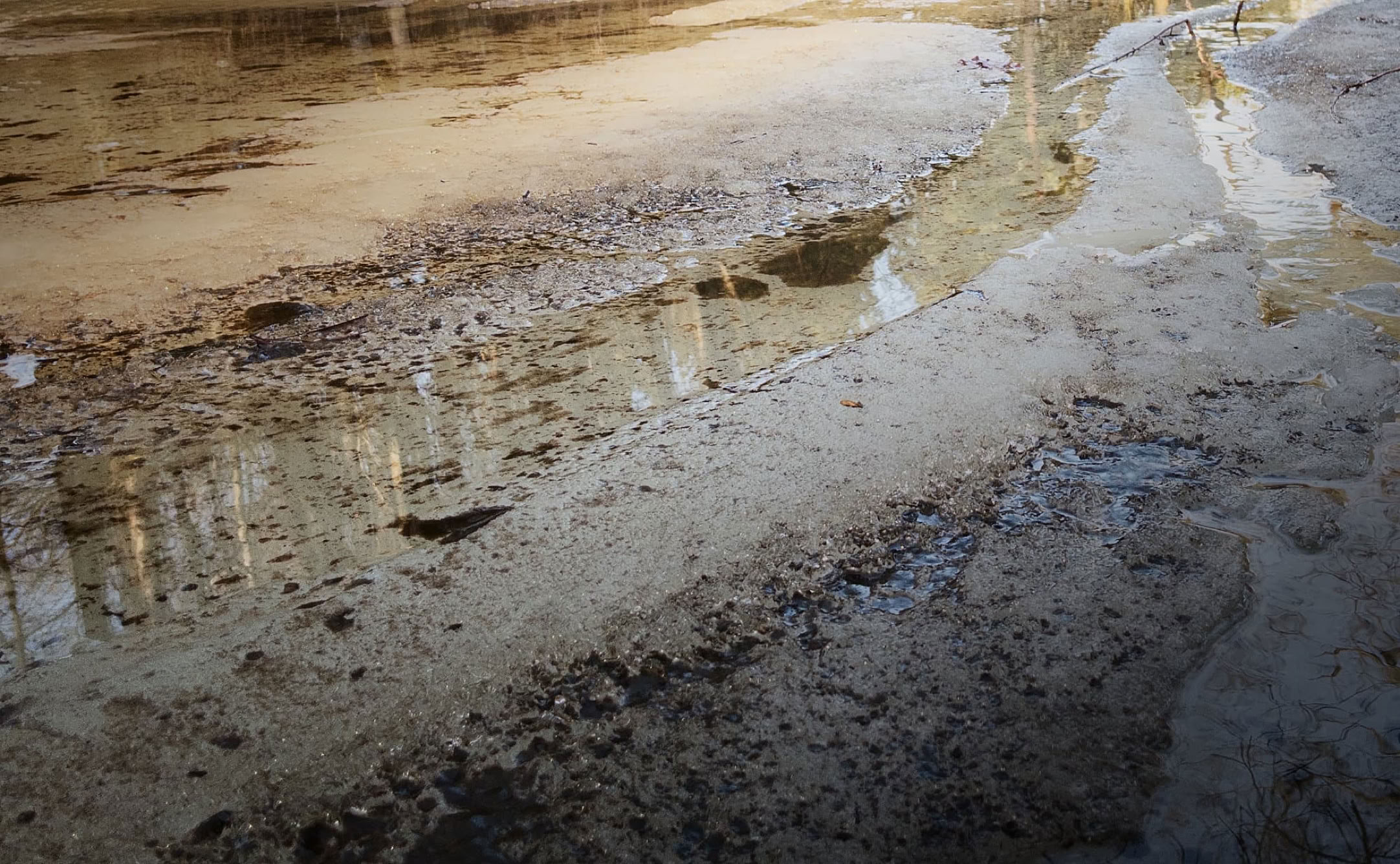 A muddy ground with puddles reflecting a building and the sky.
