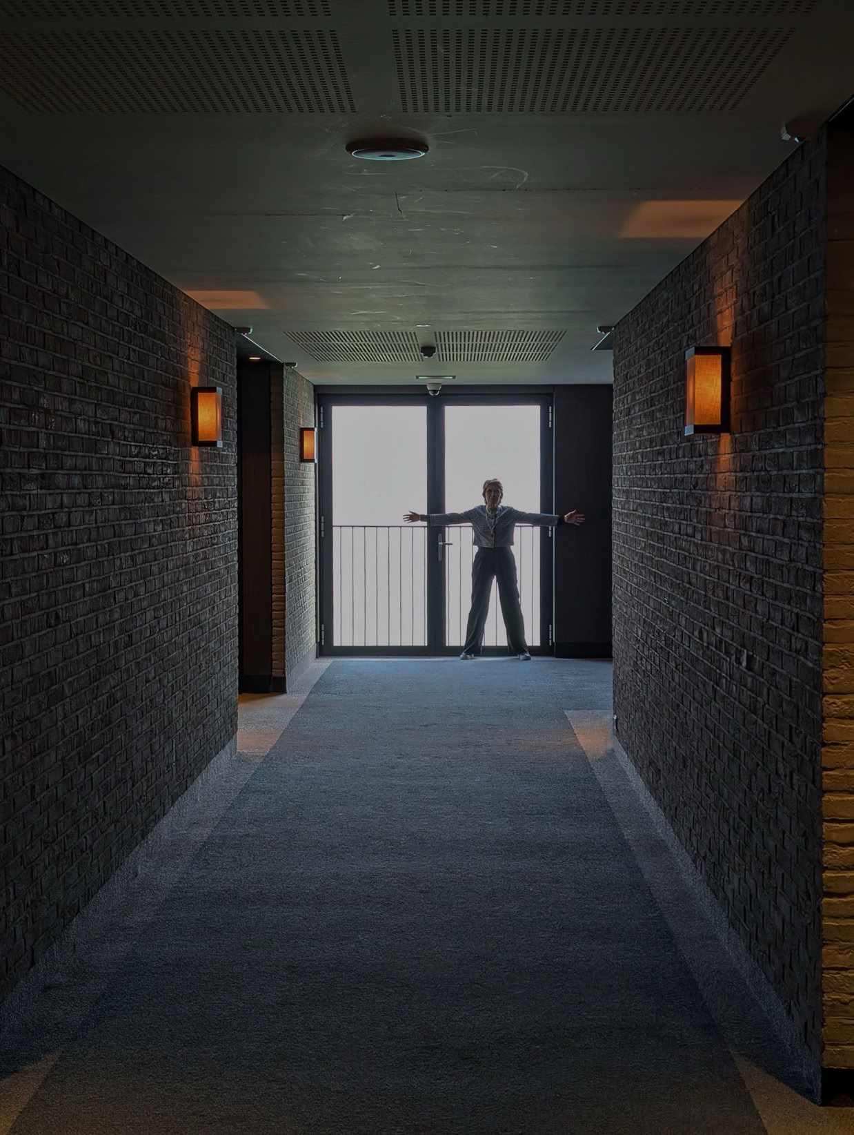 A person stands with arms outstretched in front of a large window at the end of a dimly lit hallway with brick walls and wall-mounted lights.